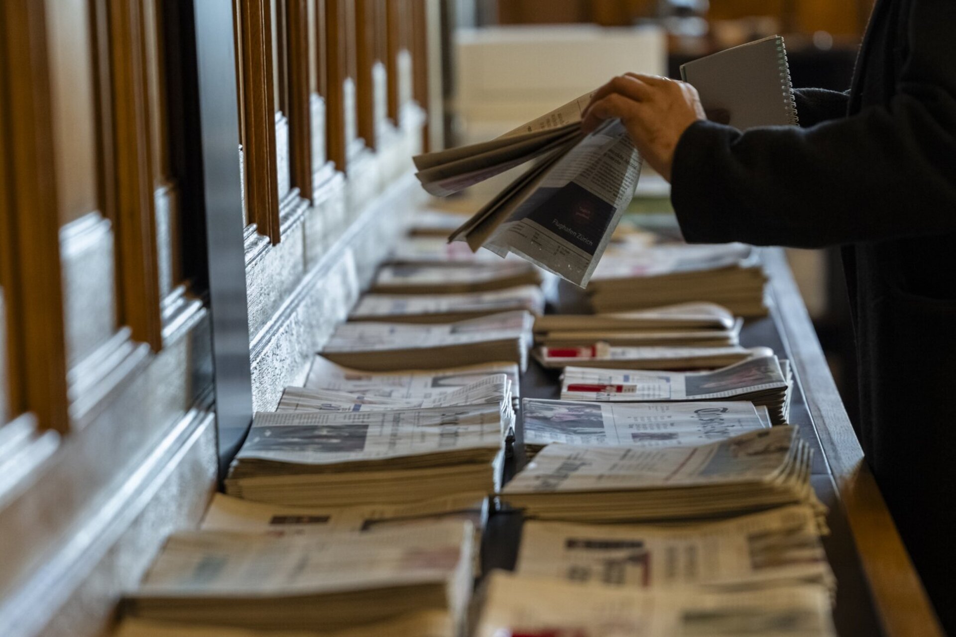Des mains feuillettent des journaux sur une table, plusieurs piles de journaux sont disposees pour consultation.