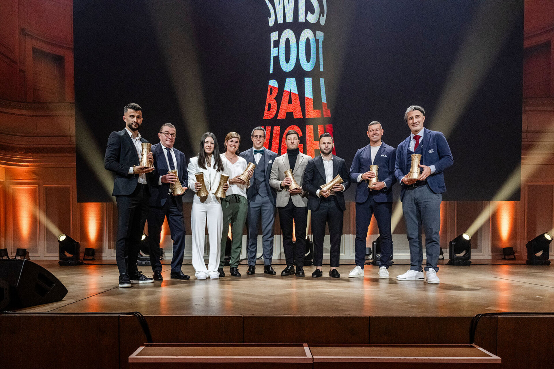 Un groupe de femmes et d'hommes debout sur la scène de la Swiss Football Night mit leurs trophées dorés devant un grand écran affichant le logo de l'événement