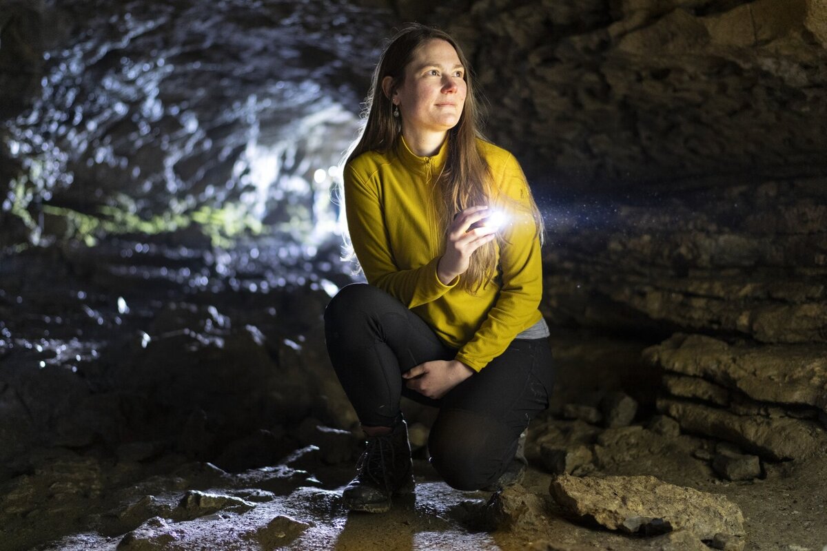 Eine Forscherin kniet im Hoehleninnern des Drachenlochs im Taminatal und leuchtet mit einer Taschenlampe die Felswaende aus.
