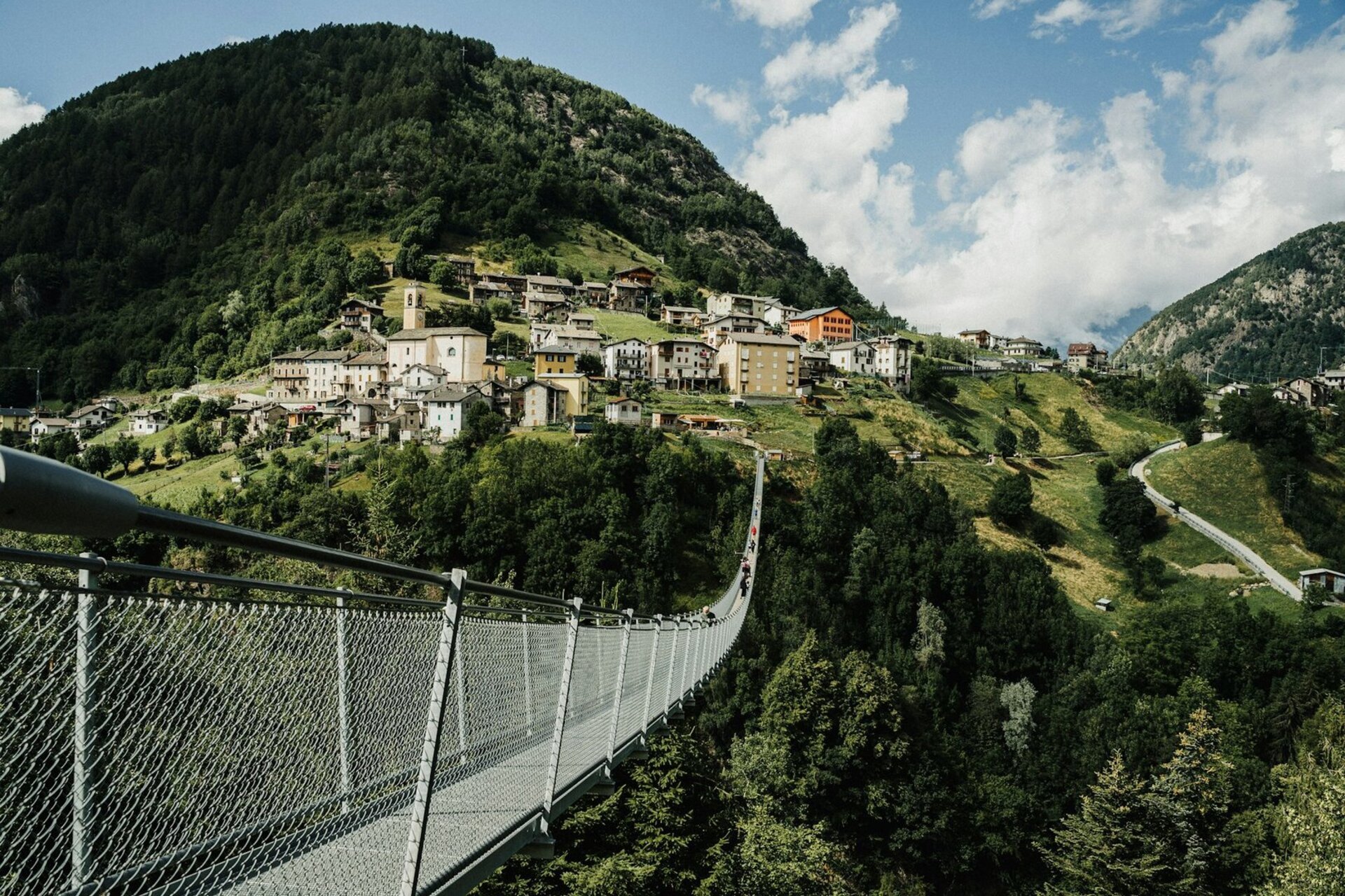 Eine schmale Hängebrücke führt über ein bewaldetes Tal zu einem kleinen Bergdorf mit dicht stehenden Häusern an einem steilen Hang, umgeben von grünen Wiesen und Bergen unter blauem Himmel mit Wolken.