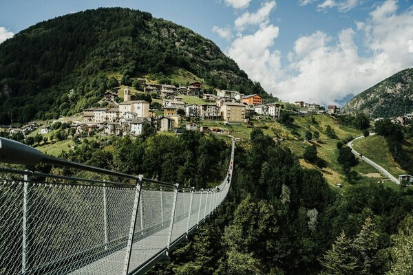 Eine schmale Hängebrücke führt über ein bewaldetes Tal zu einem kleinen Bergdorf mit dicht stehenden Häusern an einem steilen Hang, umgeben von grünen Wiesen und Bergen unter blauem Himmel mit Wolken.