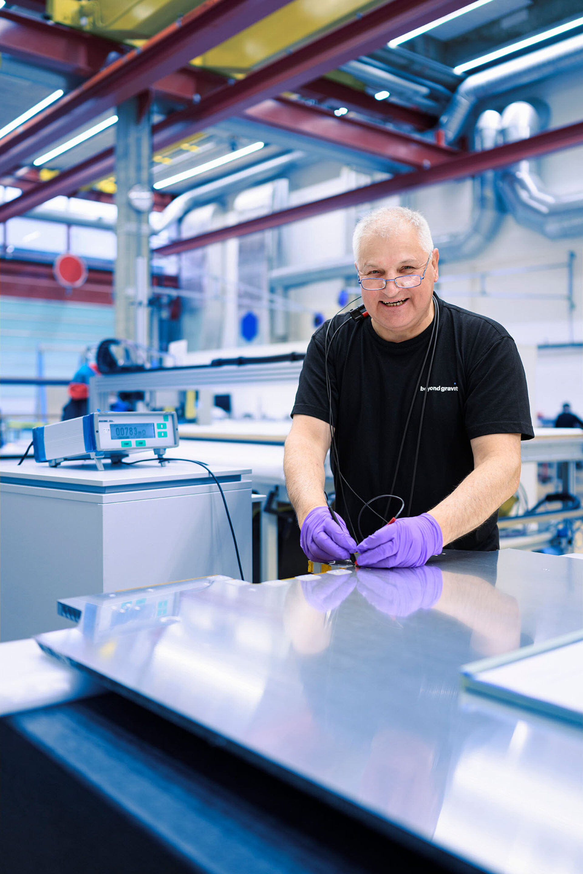 Un employé de l'entreprise Beyond Gravity portant des lunettes et des gants de protection violets effectue des mesures auf une plaque métallique brillante.