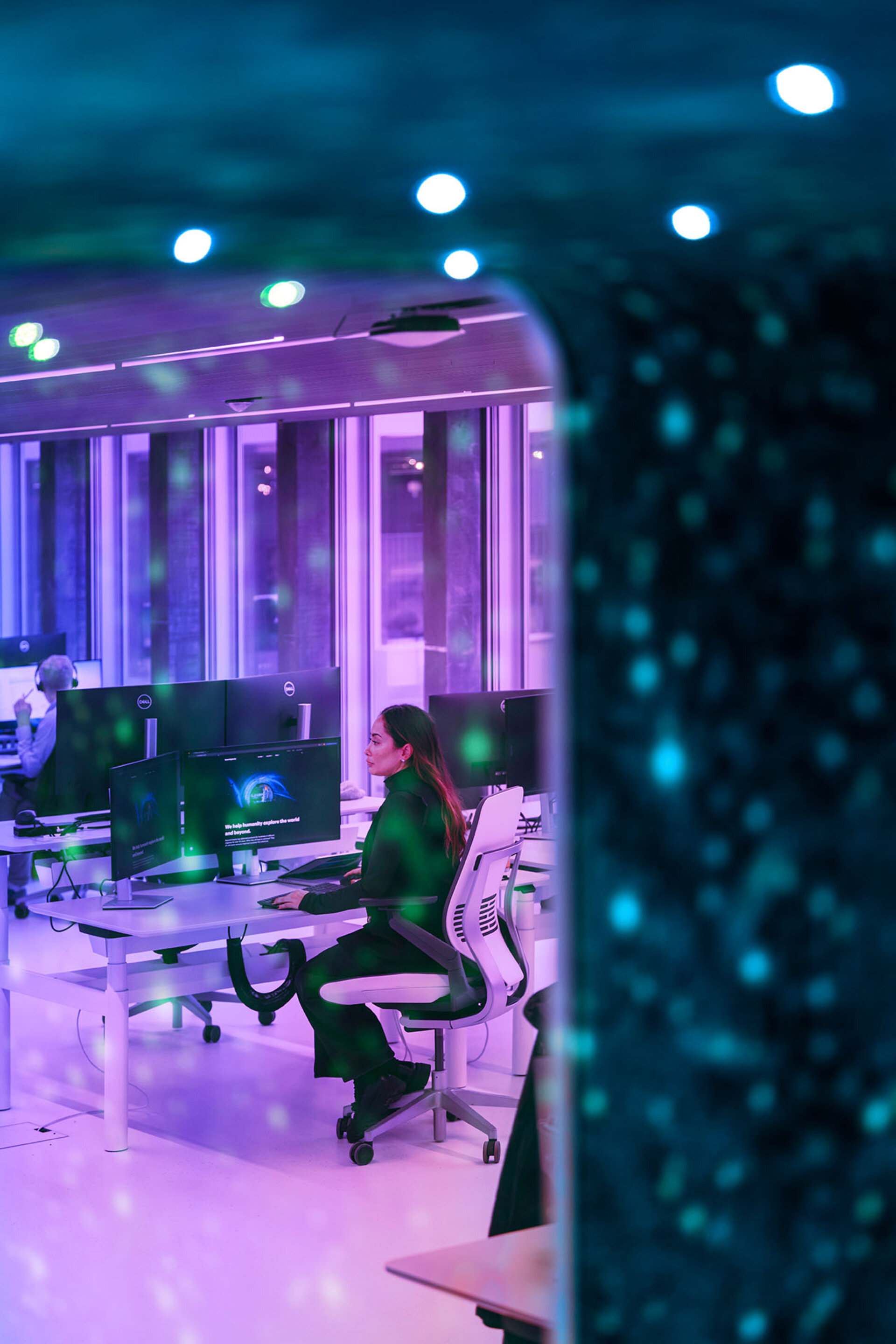 Une employée de l'entreprise Beyond Gravity travaille de manière concentrée à un poste de travail informatique moderne dans un bureau à l'éclairage violet.