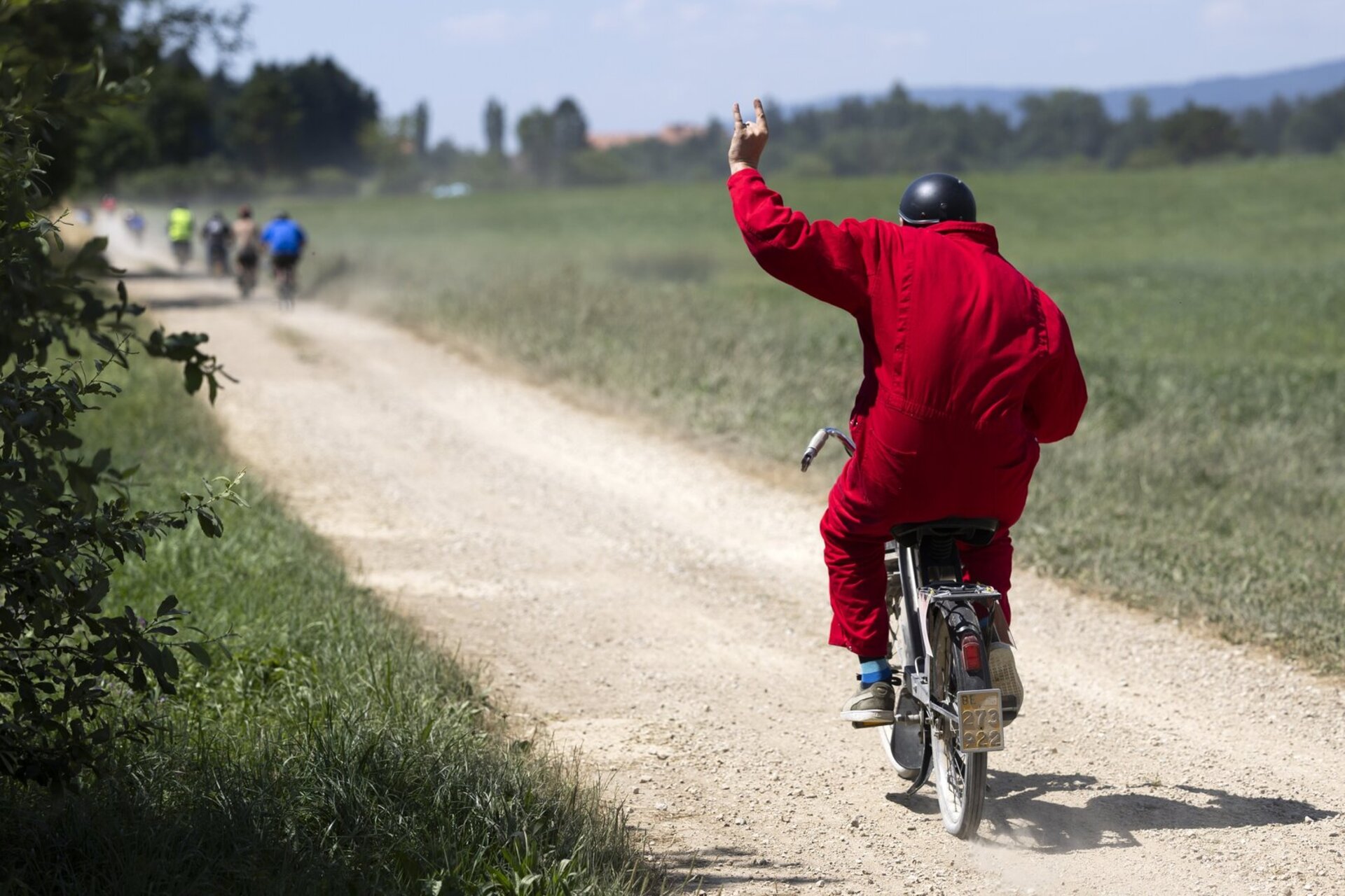ne personne en combinaison rouge roule en cyclomoteur sur un chemin de campagne et leve le bras.