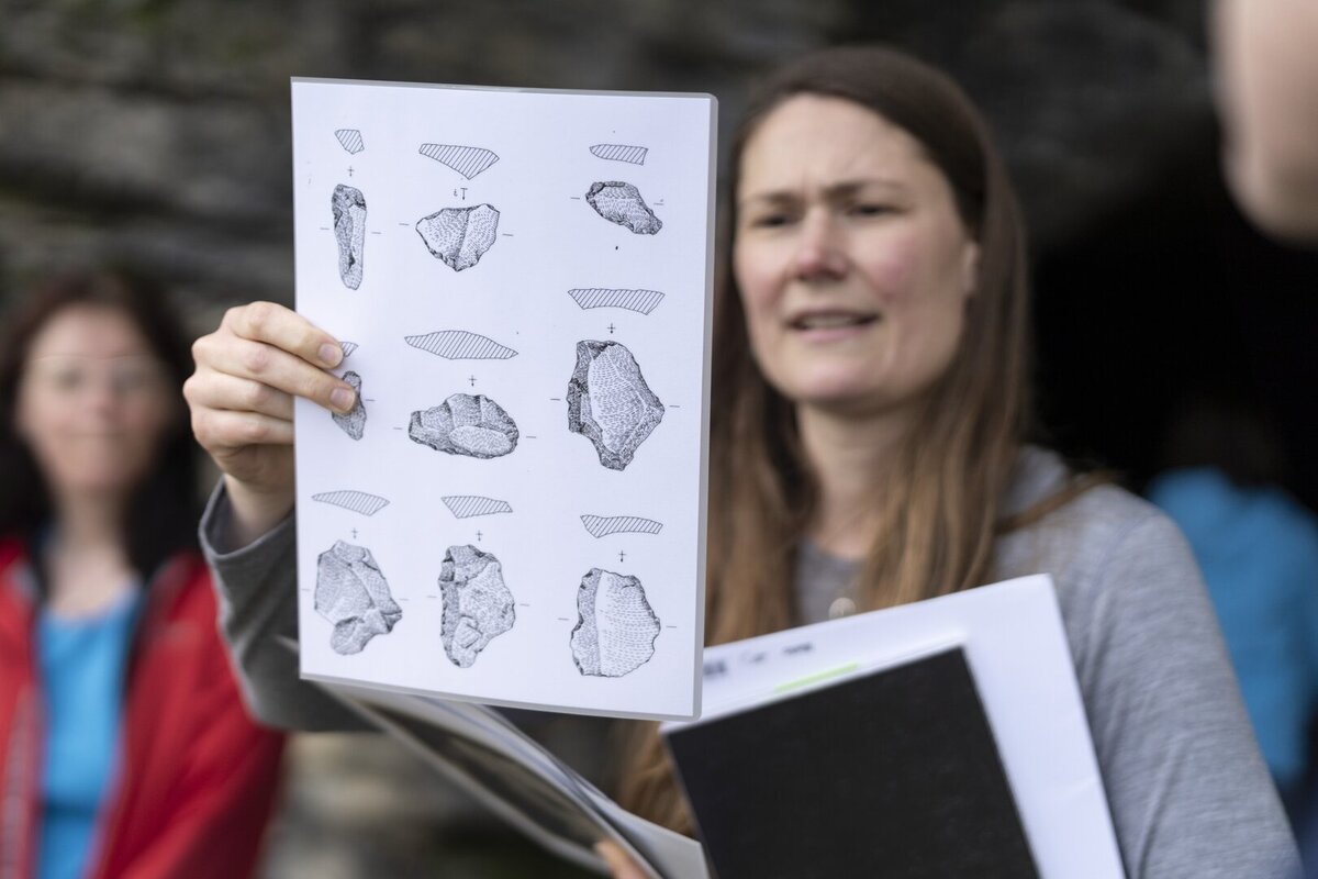 Eine Frau haelt ein Blatt mit Zeichnungen von Steinwerkzeugen hoch und erklaert die Fundstuecke vor einer Felswand.