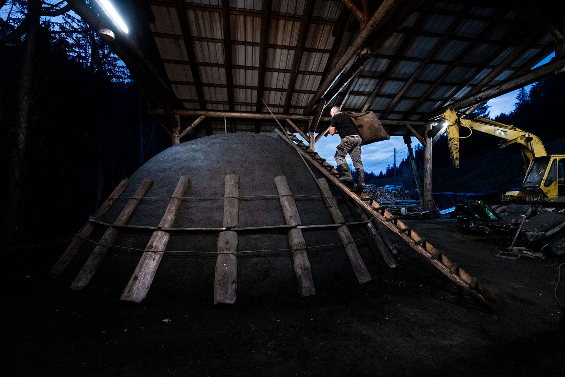 Un homme monte une echelle avec un sac de charbon de remplissage et travaille sur un grand meule de charbon sur la place a charbon de Drachslis, dans l'Entlebuch.