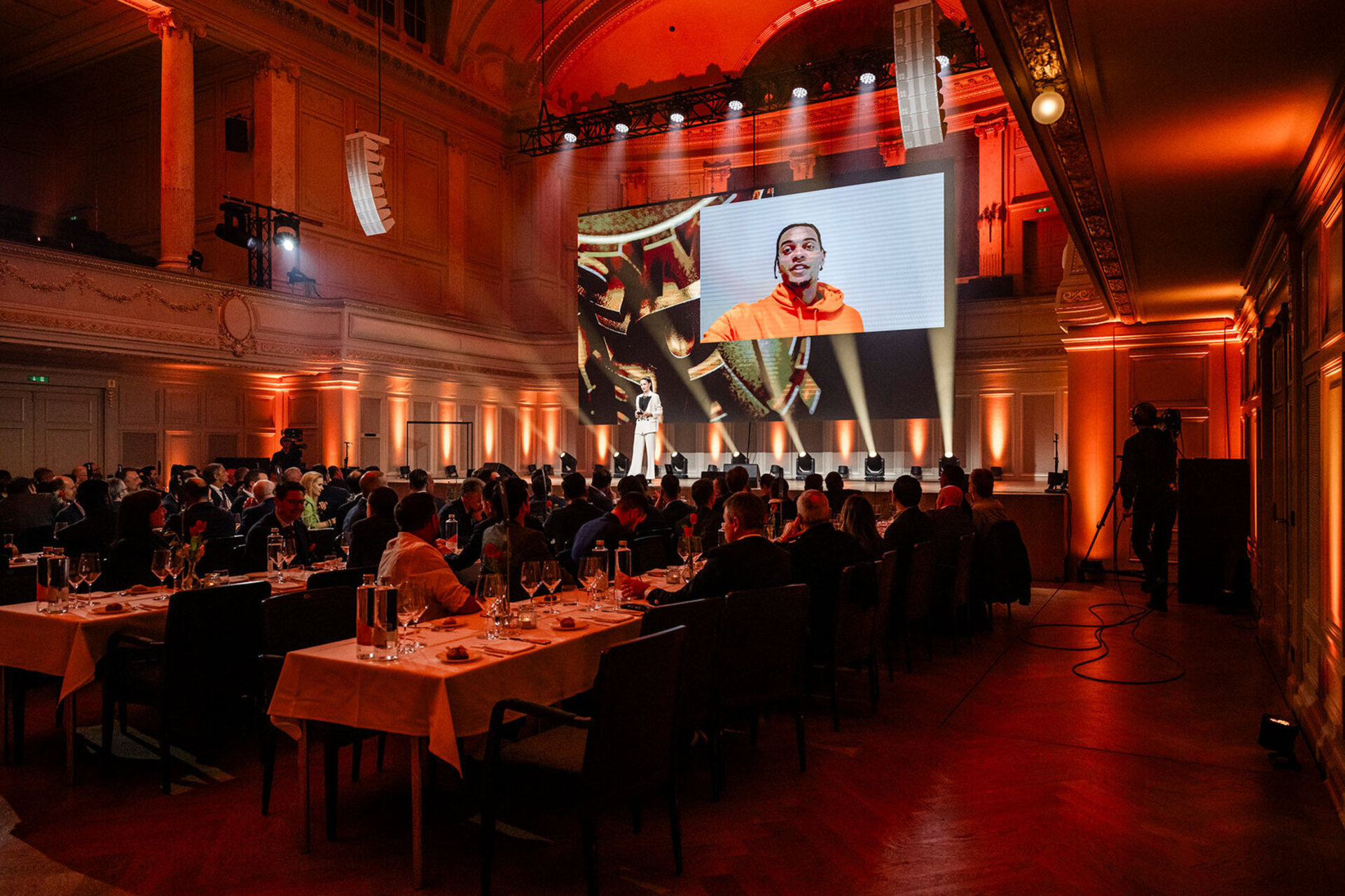 Vue de la salle de la Swiss Football Night, illuminée pour l'occasion, avec de nombreux invités attablés, tandis que Manuel Akanji apparaît par message vidéo sur un grand écran en arrière-plan.