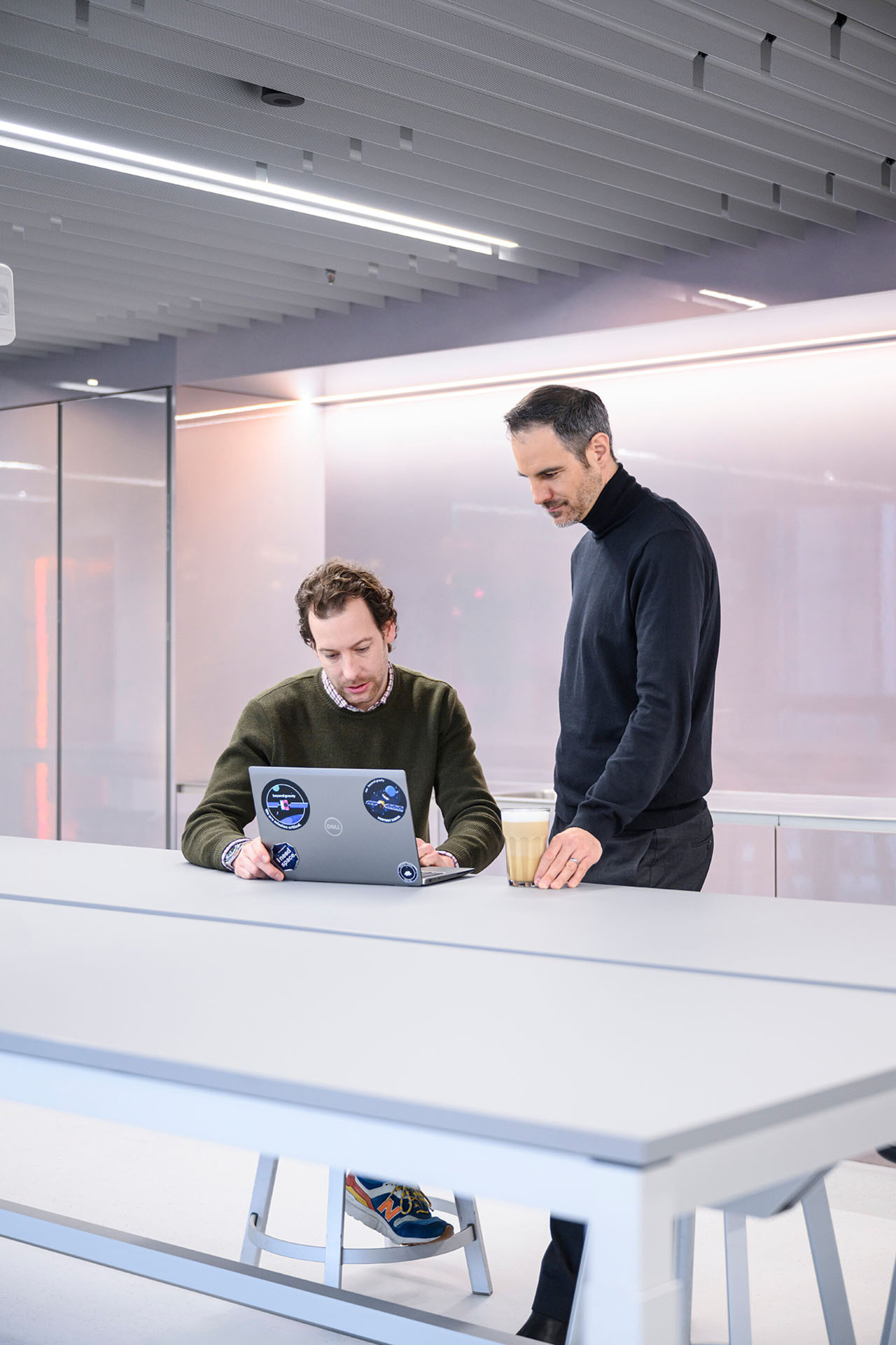 Deux hommes travaillent sur un ordinateur portable posé sur une longue table blanche dans un bureau moderne, tandis que l’un d’eux tient un verre de café.