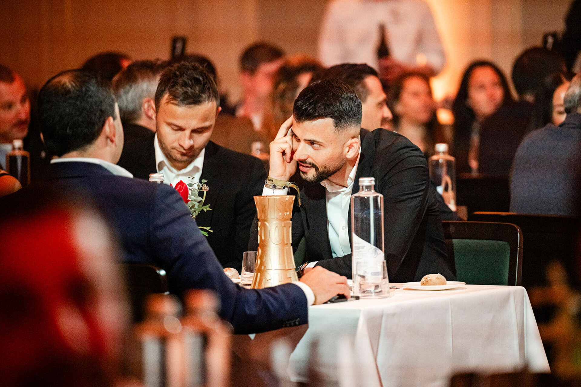 Un homme en costume sombre est assis à une table festive dans la salle de la Swiss Football Night et discute avec son vis-à-vis, tandis qu’un trophée doré est posé sur la table devant lui.