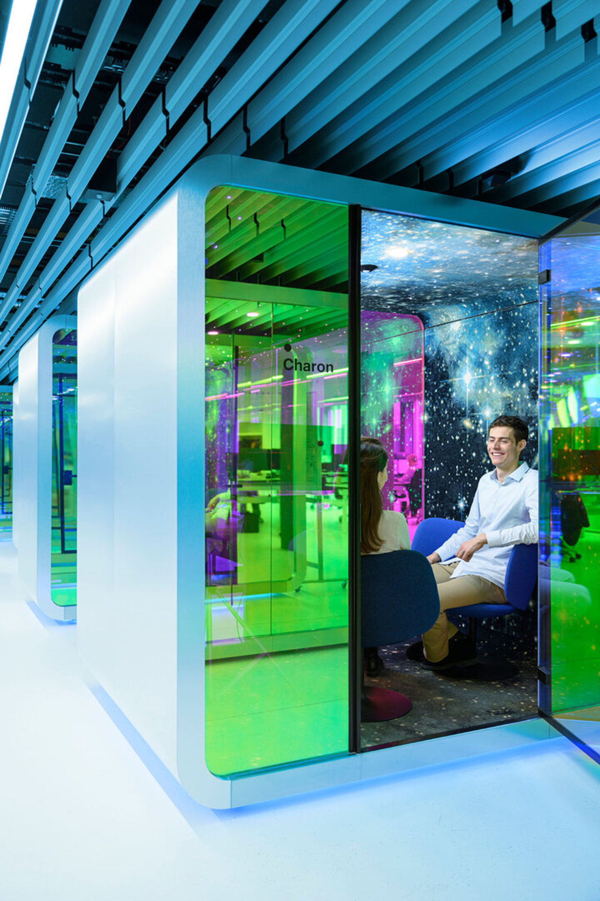 Deux personnes sont assises dans une cabine de réunion au design moderne et à l'éclairage coloré, portant l'inscription Charon, chez l'entreprise Beyond Gravity et discutent ensemble.