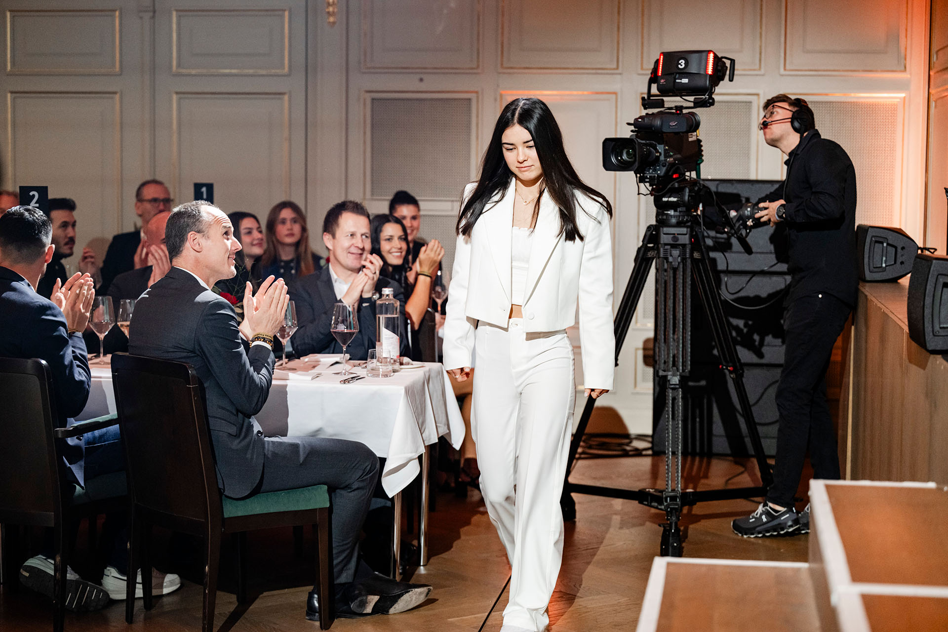 Une jeune femme en tailleur-pantalon blanc passe devant des invités qui applaudissent et une caméra de cinéma lors de la Swiss Football Night pour Keystone-ATS.