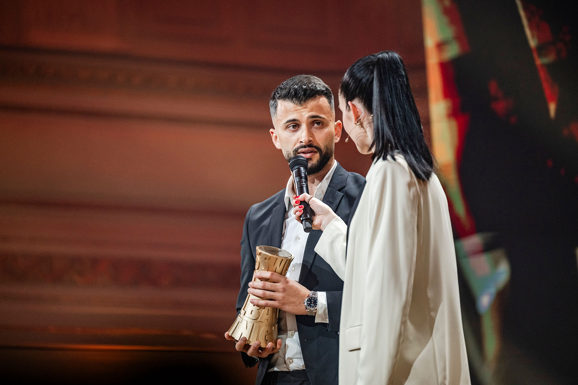 n homme en costume sombre tient un trophée doré de la Swiss Football Night dans ses mains et parle dans un micro tenu par une femme en veste claire.