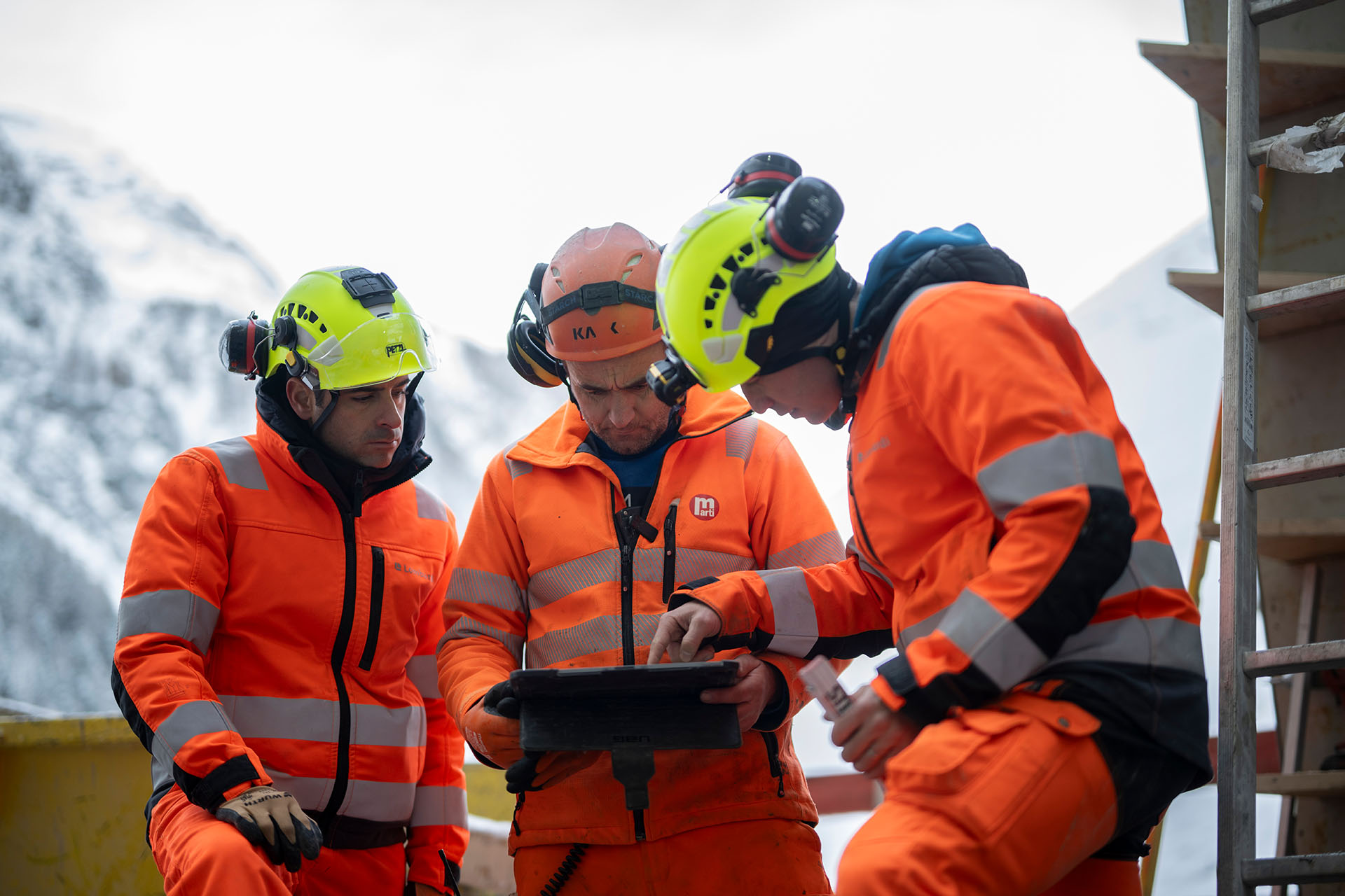 Trois ouvriers de chantier en tenue de signalisation orange et portant des casques de protection se tiennent ensemble sur le chantier du barrage de Gigerwald d'Axpo et consultent ensemble une tablette.