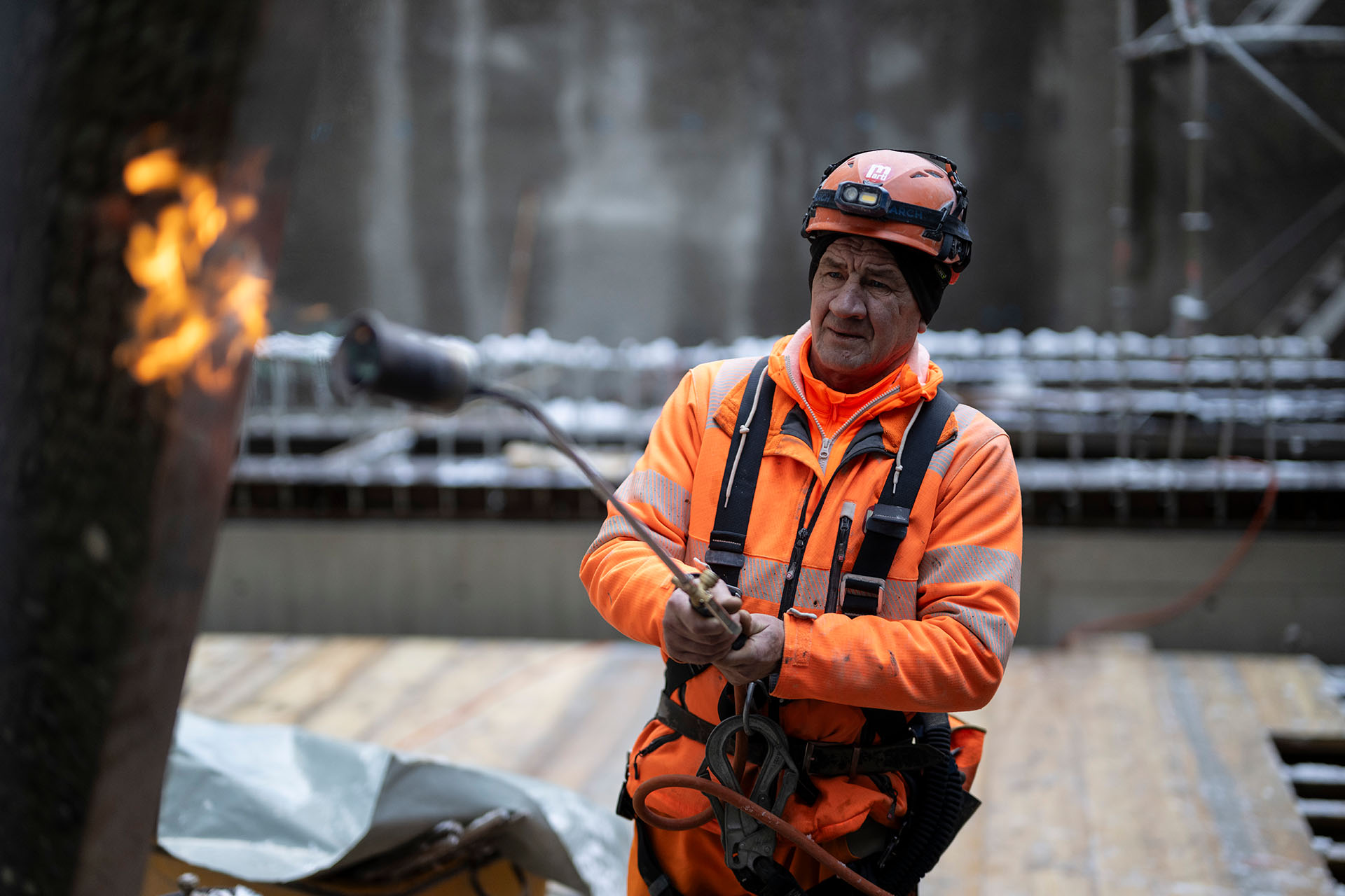 Homme en tenue de protection orange et casque, travaille sur un chantier avec un chalumeau, flamme a gauche dans l'image.