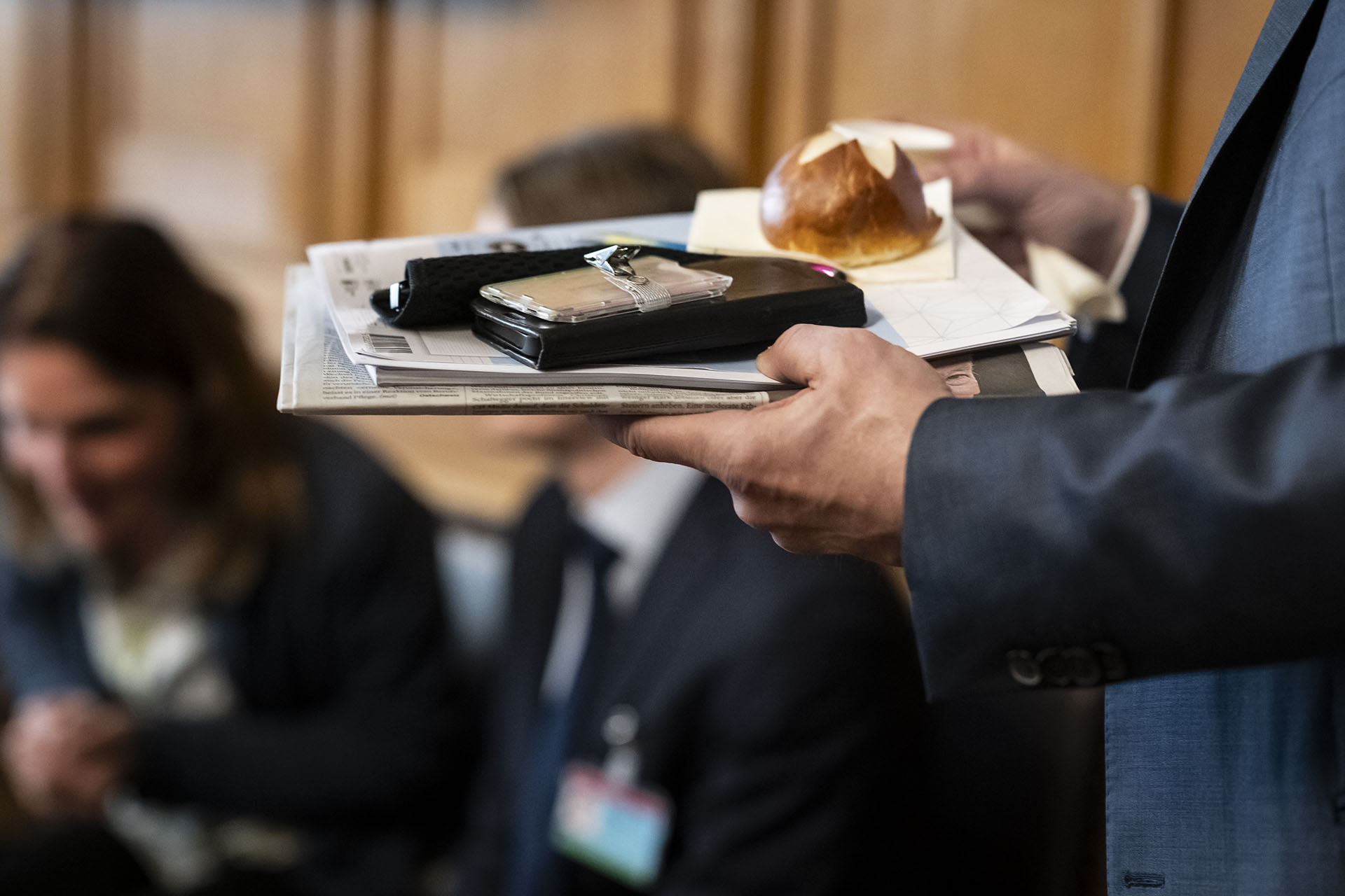 Une personne en costume bleu tenant une pile de journaux et de documents sur lesquels sont posés un smartphone et un petit pain.